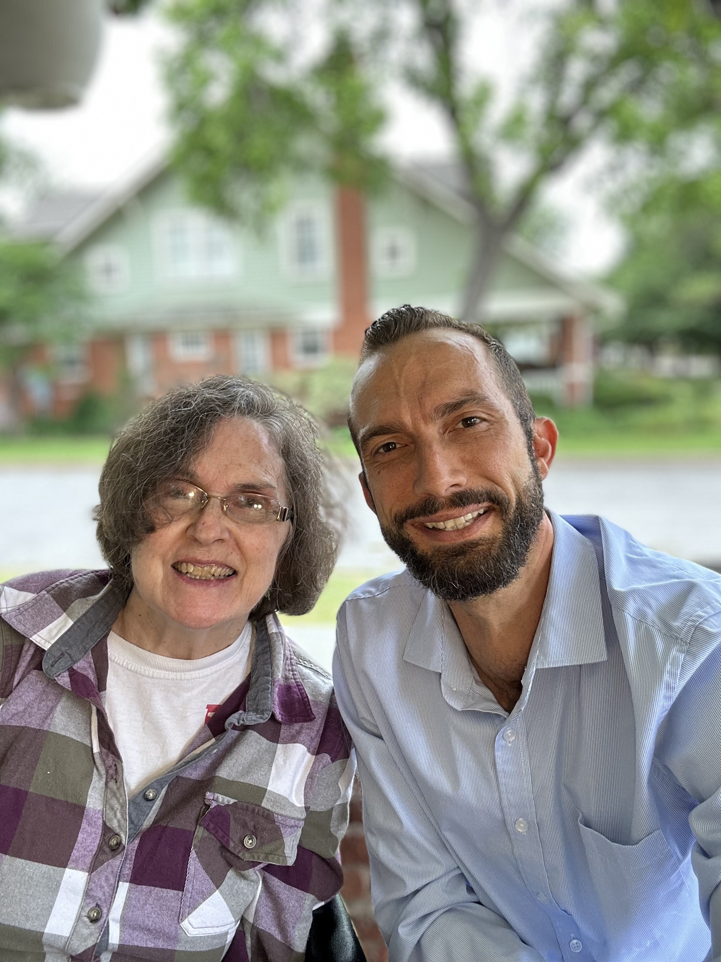 Nathan and his mother smiling warmly outdoors with a historical house blurred in the background. A heartfelt moment showing love, caregiving, and family connection.